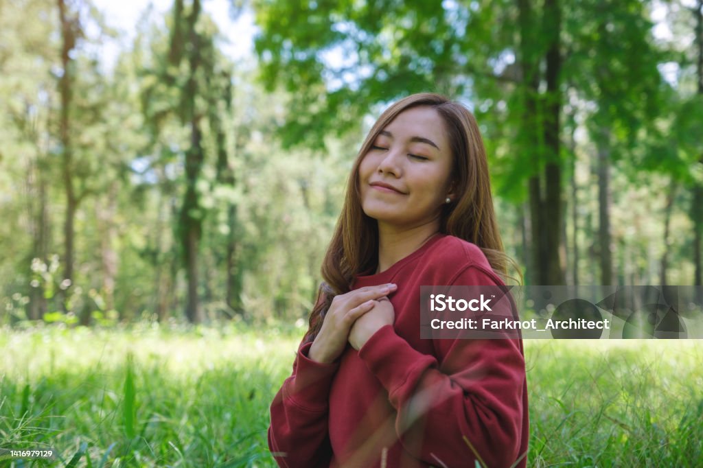 Portrait image of a young woman with closed eyes putting hands on her chest in the park Gratitude Stock Photo