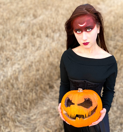Halloween portrait of a young girl wearing long black dress with open shoulders and neck with a bright makeup who holds big orange pumpkin in her hands with a field background for copy space
