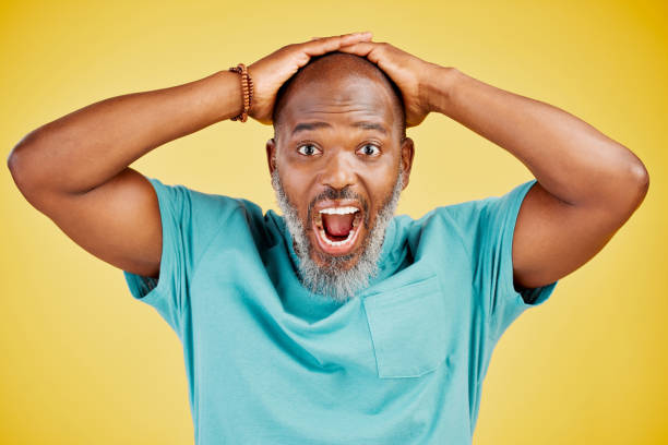 istock Mature African american man with grey beard standing with his hands on his head and looking shocked or surprised. Man with astonished facial expression while standing against yellow studio background 1416982569