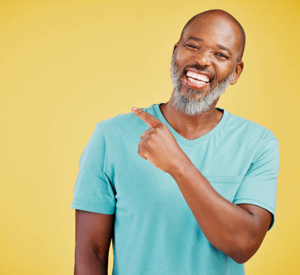 istock Mature african man laughing and pointing in a direction against a yellow studio background. Black guy giggling and making a pointing gesture reacting with laughter while looking cheerful and happy. Lighten the mood with an amusing and funny joke 1416992513