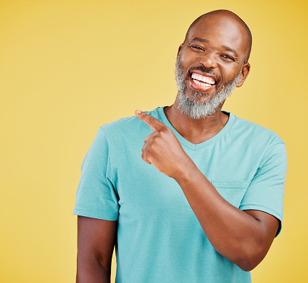 Mature african man laughing and pointing in a direction against a yellow studio background. Black guy giggling and making a pointing gesture reacting with laughter while looking cheerful and happy. Lighten the mood with an amusing and funny joke