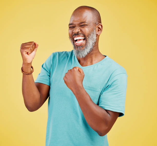 istock A mature african man looking ecstatic while while celebrating by making a fist pump gesture with his hands and cheering against a yellow studio background. Success and Celebrations infuse life with passion and purpose 1416992518