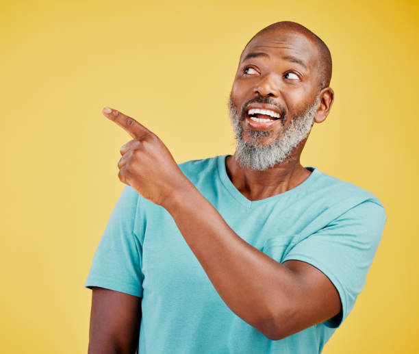 istock Mature african man smiling and pointing in a direction against a yellow studio background. Black guy looking happy and making a pointing gesture reacting with smile while looking cheerful and happy. Stay positive you never know what's coming next 1416992520