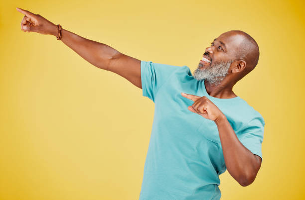 istock Mature african man smiling and pointing in a direction against a yellow studio background. Black guy looking happy and making a pointing gesture reacting with a smile while looking cheerful and happy. Stay positive you never know what's coming next 1416992521