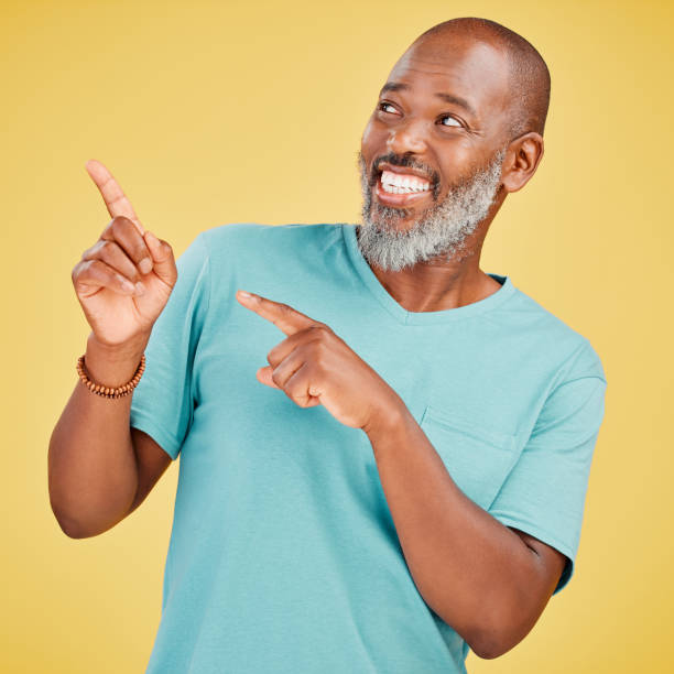 istock Mature african man smiling and pointing in a direction against a yellow studio background. Black guy looking happy and making a pointing gesture reacting with a smile while looking cheerful and happy. Stay positive you never know what's coming next 1416992526