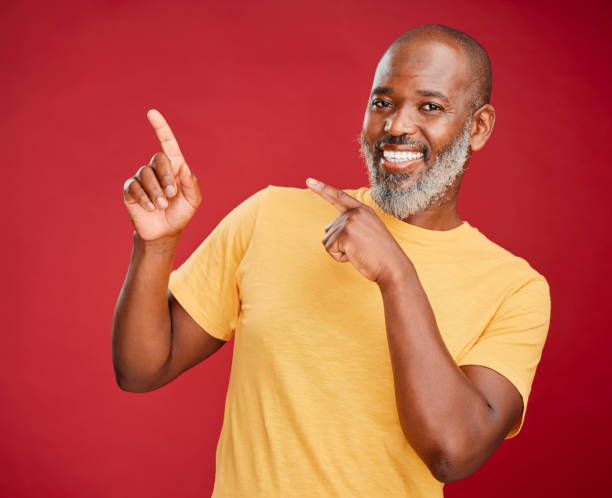 istock Mature african man smiling and pointing in a direction against a red studio background. Black guy looking happy and making a pointing gesture reacting with a smile while looking cheerful and happy. Stay positive you never know what's coming next 1416992541