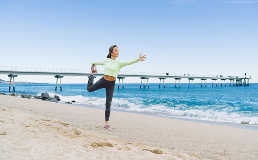Full body middle aged female in activewear standing on sandy coast near blue sea water while doing lord of dance pose