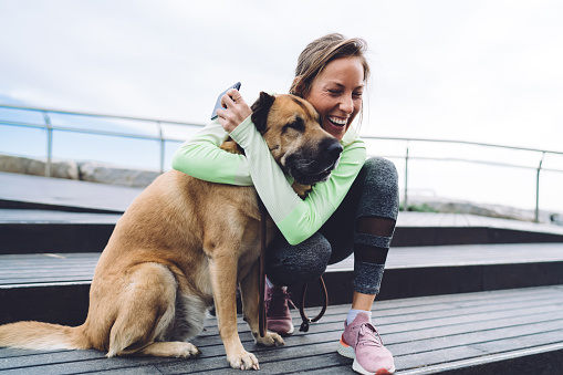 Cheerful mature female in activewear laughing and hugging dog while sitting on stone stair and holding mobile phone in hand