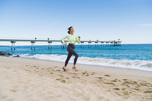 Full body smiling adult female athlete in sportswear doing cardio during workout running along seaside near waving blue sea in sunny day