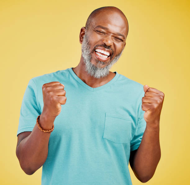 istock A mature african man looking ecstatic while while celebrating by making a fist pump gesture with his hands and cheering against a yellow studio background. Success and Celebrations infuse life with passion and purpose 1416996478