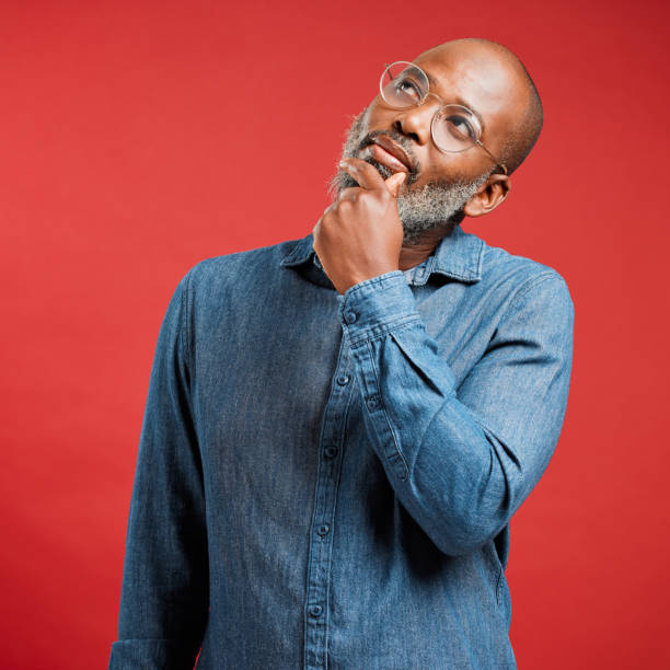 un homme afro-américain mature portant des lunettes. isolé sur un fond de studio rouge. homme noir rêvant et pensant tout en frottant sa barbe grise et en ayant l’air conscient - raised eyebrows photos photos et images de collection