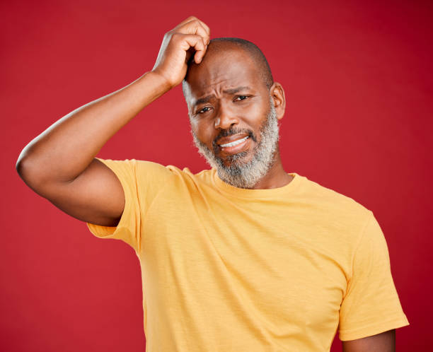 Mature african american man with a beard scratching his head standing against a red studio background. Unhappy, frowning, unsure. Anxiety, sad, worried Mature african american man with a beard scratching his head standing against a red studio background. Unhappy, frowning, unsure. Anxiety, sad, worried confusion raised eyebrows human face men stock pictures, royalty-free photos & images