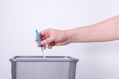 A hand tosses a used syringe in the trash on a gray background. The concept of medical waste disposal.