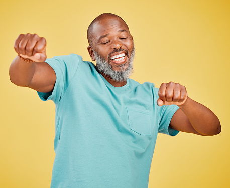 A mature african man looking ecstatic while while celebrating and dancing by making a fist pump gesture with his hands and singing against a yellow studio background. Dance like nobody's watching