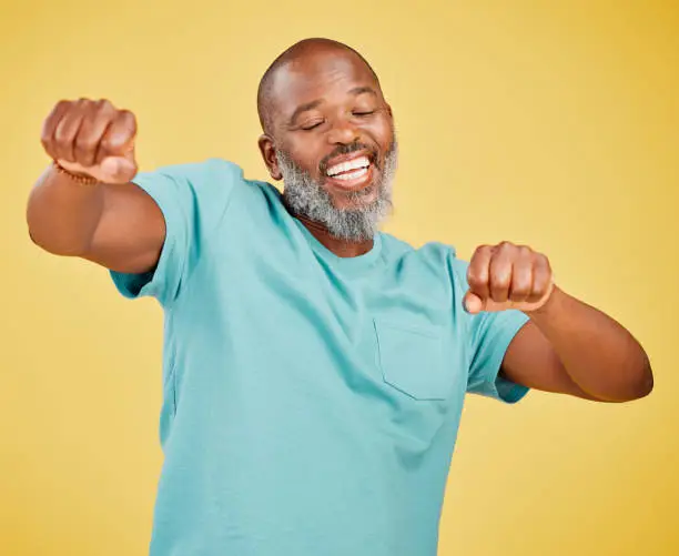 Photo of A mature african man looking ecstatic while while celebrating and dancing by making a fist pump gesture with his hands and singing against a yellow studio background. Dance like nobody's watching