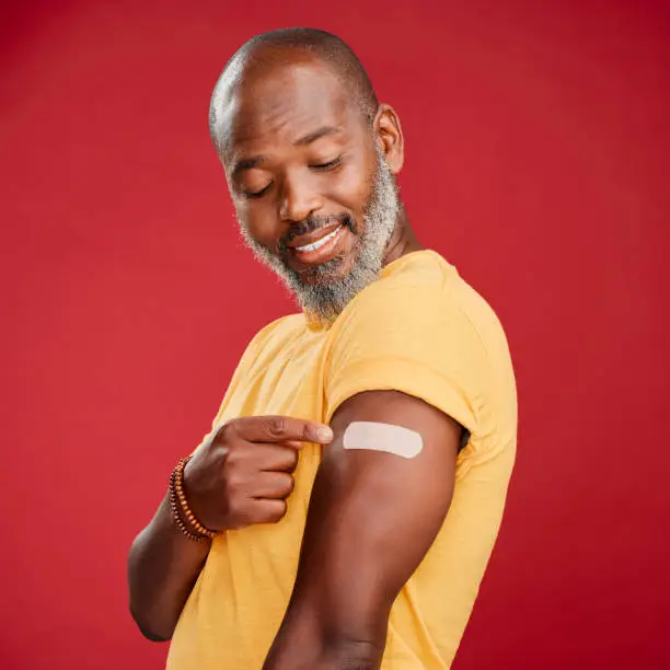 Photo of A mature african american male pointing at a plaster on his arm against a red studio background. Black african man smiling after a minor injury and  making a hand gesture towards the plaster on his arm.