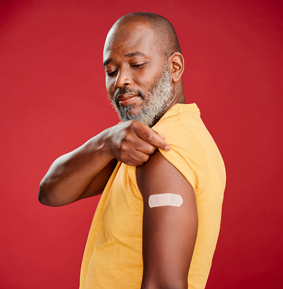 Mature african american man with a beard smiling and looking happy while wearing a bandaid and standing against a red studio background. Vaccination, health and healthcare is important to cure the virus