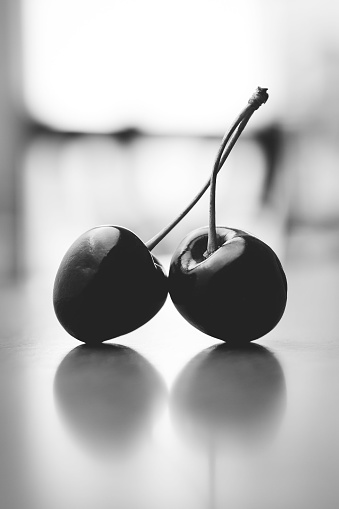 A vertical shot of two cherries tied together and reflected on a white surface, greyscale shot