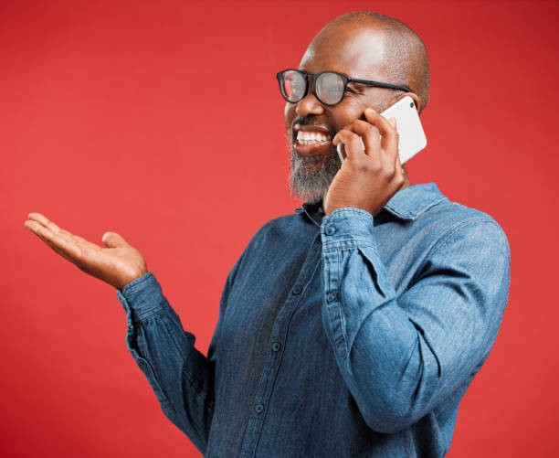 istock Happy mature African American man isolated against a red background in studio, using hand gestures to ask questions while talking on a cellphone. Cheerful black man with glasses chatting on a phone 1417021160