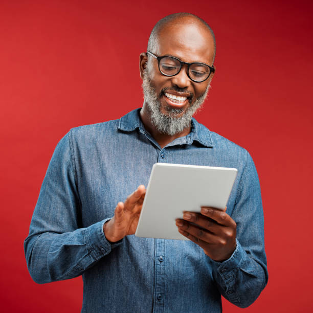 smiling man browsing on a tablet online, networking on the internet and scrolling on an app while standing against a red studio background. happy, mature and african male reading an email and typing - male african descent africa ethnic imagens e fotografias de stock