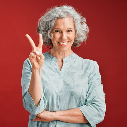 La mujer mayor expresa el signo de paz, el gesto v y el emoji con los dedos sobre el fondo rojo del estudio. Retrato de una dama despreocupada, fresca y sonriente en un estado de ánimo positivo, juguetón y divertido que muestra la victoria con la mano photo