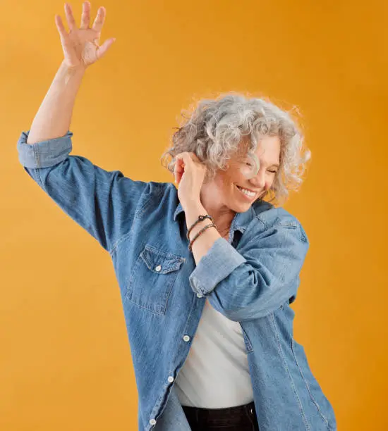 Photo of Celebrating, partying and dancing mature woman, happy and cheerful senior making waving hand gesture and smiling. Elderly caucasian woman having fun while she dances against an orange background