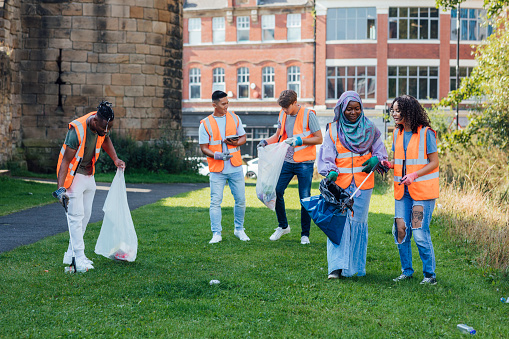 A group of friends who care for the environment, wearing reflective vests and using litter pickers and plastic bags to pick up litter on a grass area in the centre of Newcastle, North East England. They are bonding while they help clean up the community.