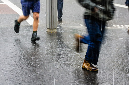 Pedestrians rush to avoid heavy rain in a city street, with water on the road and sidewalk and the raindrops splashing all around