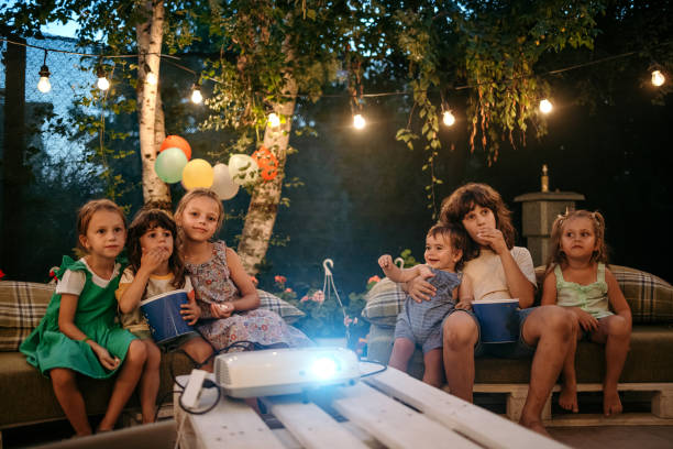 Let's watch our favorite cartoon! Group of children are sitting in the yard during a birthday party and watching a movie on a video projector happy birthday cousin images stock pictures, royalty-free photos & images