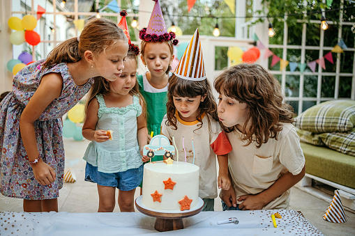 Children with cake standing around table on birthday party in garden in summer.