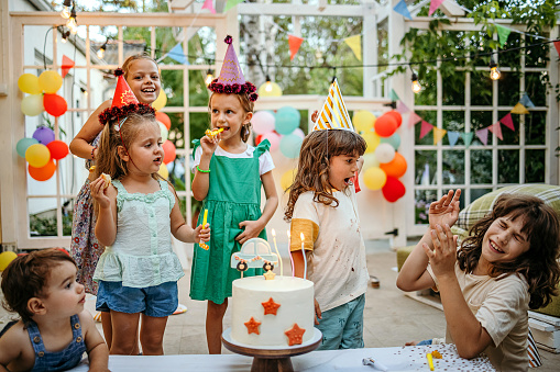 Children with cake standing around table on birthday party in garden in summer.