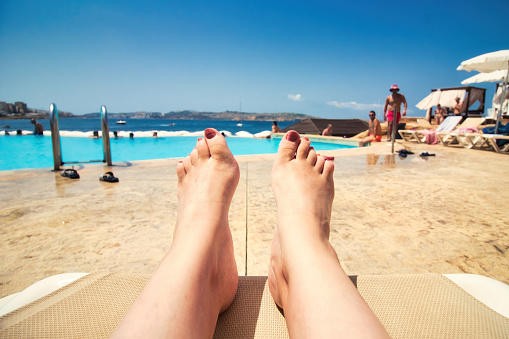 First-person point of view from a woman lying relaxed on a poolside sun lounger looking at the swimming pool with people in the background and copy-space for text