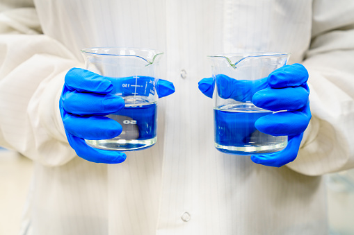A person in a lab coat is holding two beakers with clear liquids