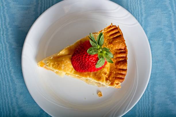 Strawberry lover’s dessert with fresh whole strawberry garnish covering a flaky apple pie on a white plate set on blue tabletop. Top down view