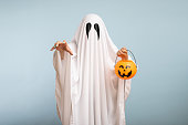 Halloween Concept. A white ghost with black eyes, made from a bedsheet with pumpkin basket for candy on blue background