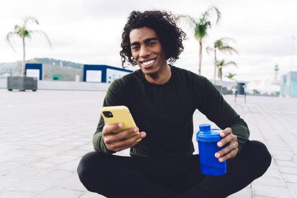 istock Cheerful sportsman using smartphone while sitting on paved street 1417064238
