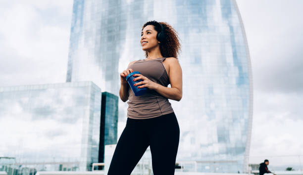 smiling sportswoman with water bottle standing near glass skyscraper - exercising motivation looking up african descent imagens e fotografias de stock