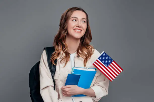 Happy girl student holding backpack, book, notebook, passport and USA flag isolated on a dark grey background, copy space