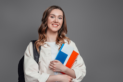 Happy girl student holding backpack, book, notebook, passport isolated on a dark grey background, copy space, immigration, paperwork