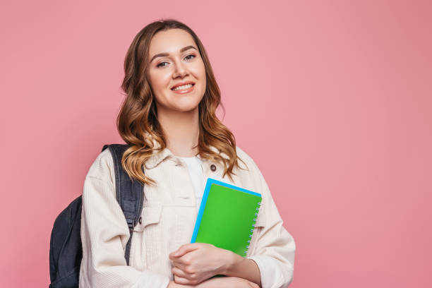 istock Happy girl student with backpack smiles and holds a notebook isolated on pink background, copy space, web banner. The concept of education, learn English 1417072003