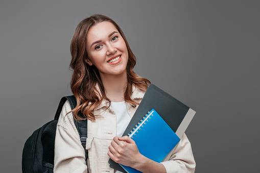 Happy girl student in bright clothes holding a notebook, notepad, a folder in her hands, smiling and looking at the camera on a dark grey background, copy space for text
