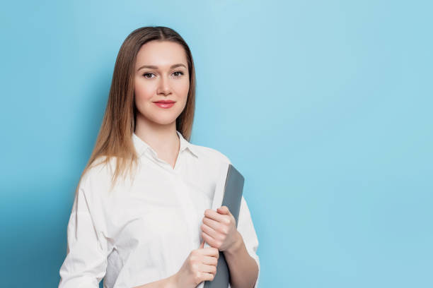 istock student girl in white clothes holding a notebook, notepad, a folder, smiling and looking at the camera isolated over blue background, copy space for text 1417073193