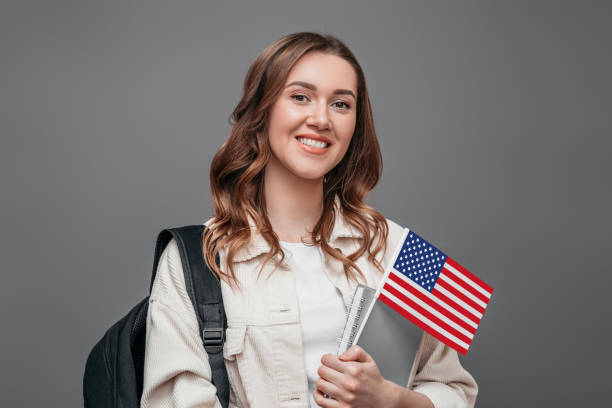 istock Girl student smiling, holding backpack and USA flag isolated on grey background, copy space, student exchange concept. Portrait of a cute student girl on a dark background with the flag of America 1417073321