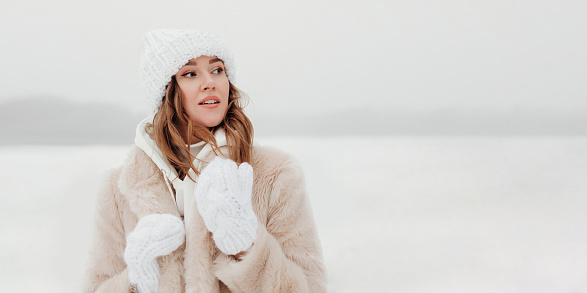 Young caucasian woman in a fur coat, white hat and gloves looking to the side against a background of snow