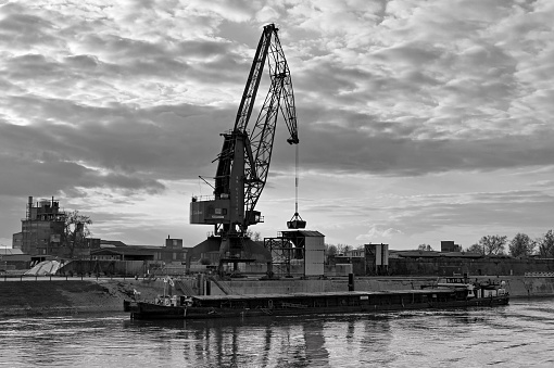Mannheim, Germany - April 2022: Silhouette of a crane unloading an industrial barge on the River Rhine at dusk. Monochrome.