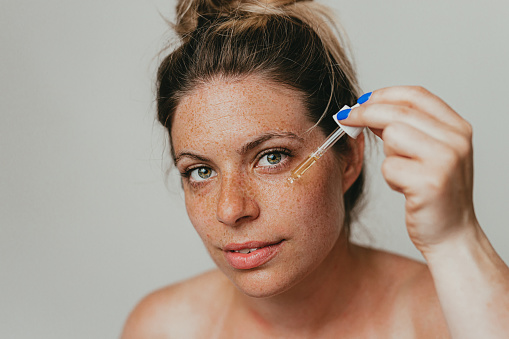 Portrait of a beautiful young freckled woman applying face serum, studio shoot in front of a white background