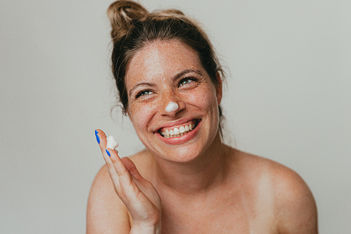 Portrait of a beautiful young freckled woman applying face cream, studio shoot in front of a white background
