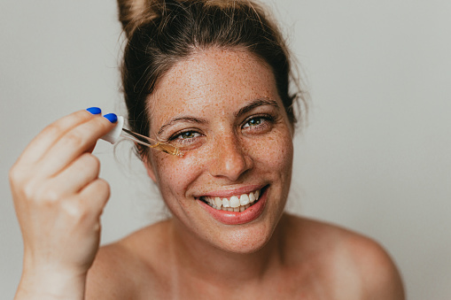 Portrait of a beautiful young freckled woman applying face serum, studio shoot in front of a white background