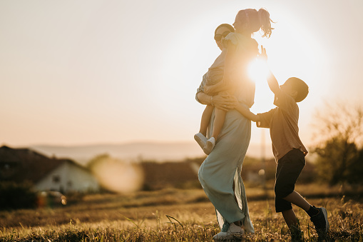 Mother taking a walk on a meadow in nature with her little daughter and son. She is carrying her little daughter in her arms.