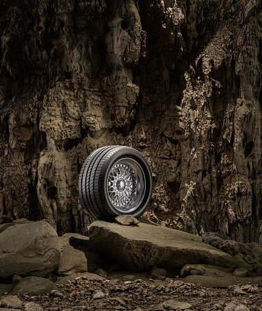 A new car wheel standing on a rocky stage. A car tire standing upright and sideways on rocky ground in front of a natural cliff. 3d rendering, no people